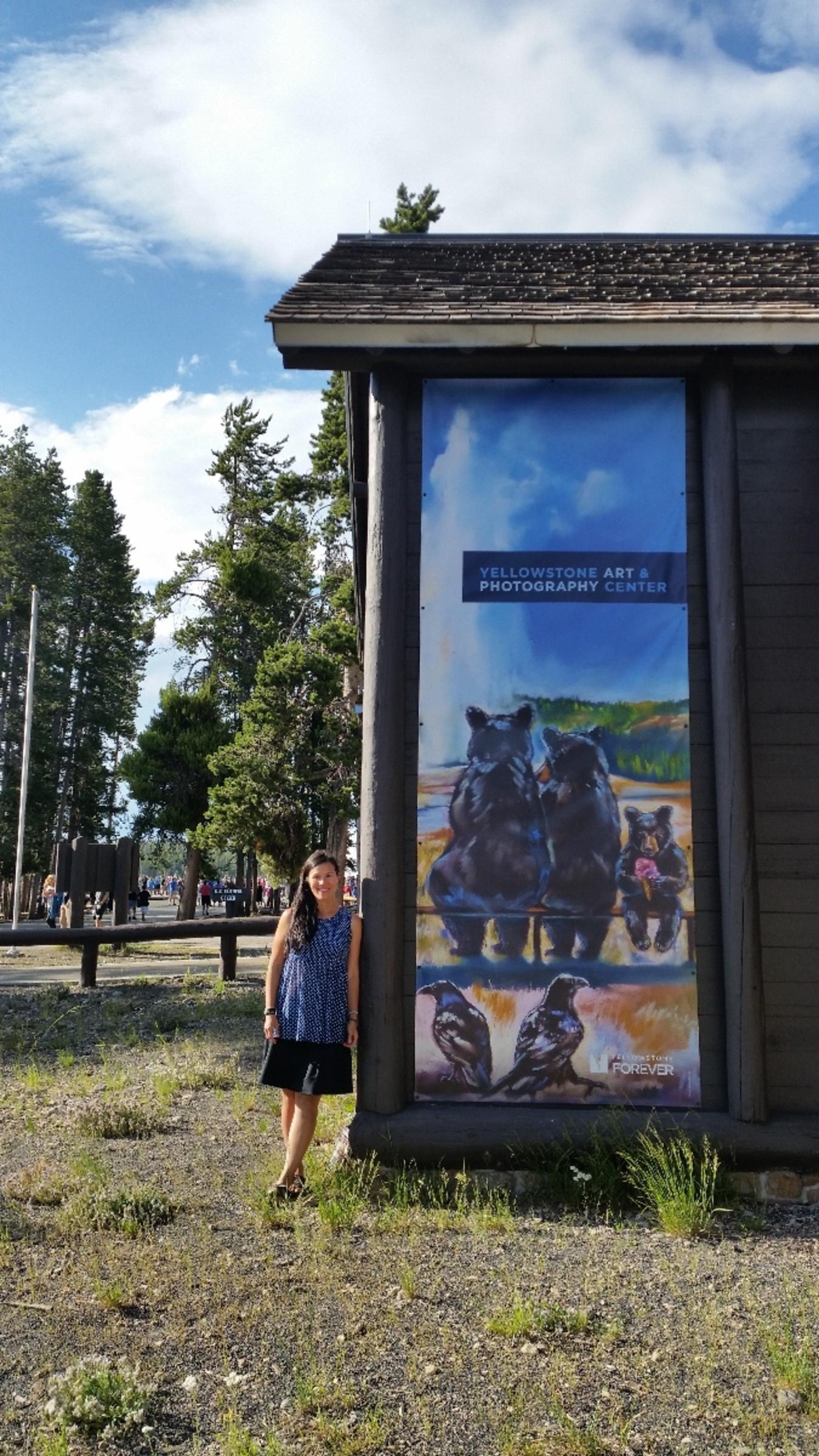 Mimi Matsuda standing next to a larger than life-sized portrayal of ravens and bears watching Old Faithful erupt.  The piece is part of an informational kiosk for Yellowstone Forever, located within eyeshot of the famous geyser.  It is a promotion for the Yellowstone Art 