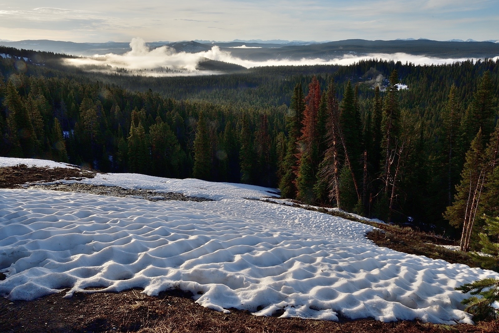The last remnants of a spring sun-cupped snow cornice overlooks steam plumes rising from geysers and hot springs scattered unto the horizon across the Yellowstone Plateau. In the distance  the Absaroka mountain range rises above the rim of the eastern horizon. Photo courtesy Steven Fuller