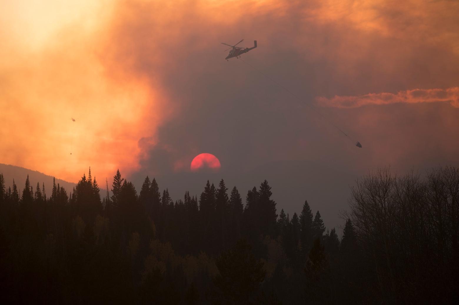 Helicopters drop water buckets against the setting sun on the 2019 Roosevelt Fire in Wyoming. Photo by Kari Greer